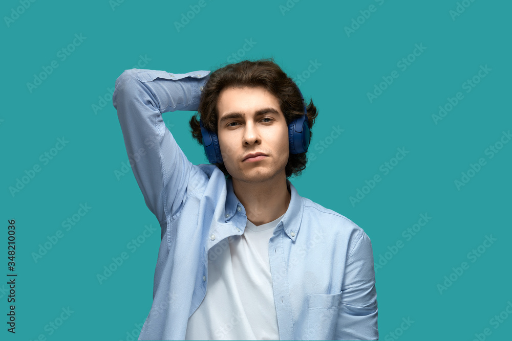 Fototapeta premium I don't care. Portrait of a young beautiful man wearing white t-shirt and blue shirt in headphones threw hand behind his head looking at camera