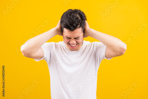 Portrait Aaian handsome young man standing wearing white t-shirt he covering his ears with hands and shouting opened mouth shriek annoyed expression, studio shot isolated yellow background