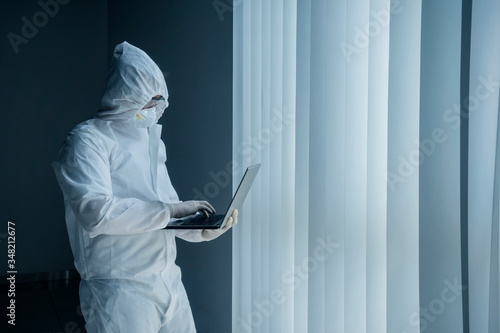 Doctor in protective suit uses laptop near window