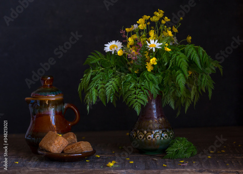 Still life with wildflowers in a retro vase on a wooden table. Nearby is a small vintage brown teapot and a plate of cakes. Black background. Bouquet of meadow flowers in a rustic jar. photo