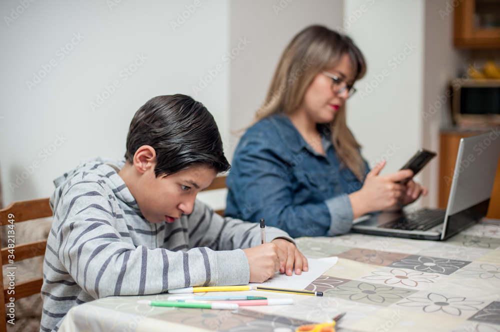 Boy studying at home while his mom is working. Focus on foreground. Boy studying at home while his mom is working. Focus on foreground.