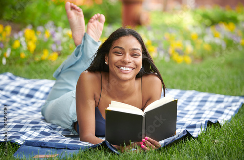 Cute young African American woman reading book on picnic blanket in shade - in front of blooming flowers photo