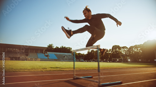 Shot of Athletic Fit Man in Grey Shirt and Shorts Hurdling and Jumping Over Barriers on a Warm Summer Afternoon in the Stadium. Athlete Doing His Routine Sports Practice.