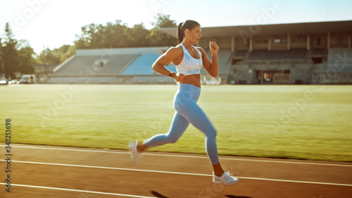 Beautiful Fitness Woman in Light Blue Athletic Top and Leggings Jogging in a Stadium. She is Running Fast on a Warm Summer Afternoon. Athlete Doing Her Routine Sports Practice. © Gorodenkoff