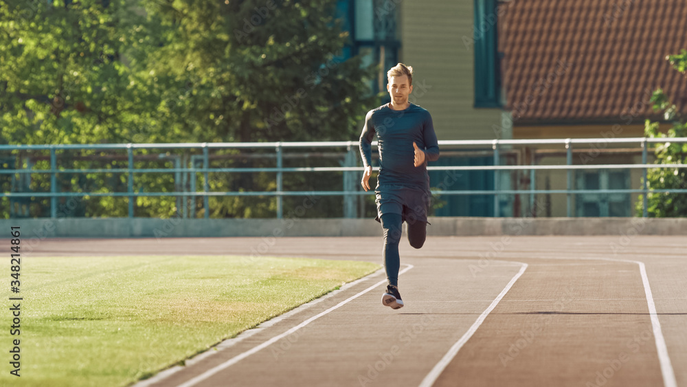 Fototapeta premium Smiling Athletic Fit Man in Grey Shirt and Shorts Jogging in the Stadium. He is Running Fast on a Warm Summer Afternoon. Athlete Doing His Routine Sports Practice.