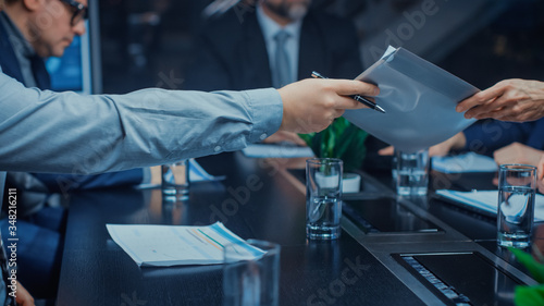 In the Corporate Meeting Room Close-up on the Hands of Businessperson Passes Folder with Important Documents. Signing Contracts Concept. People Sitting at Conference Table
