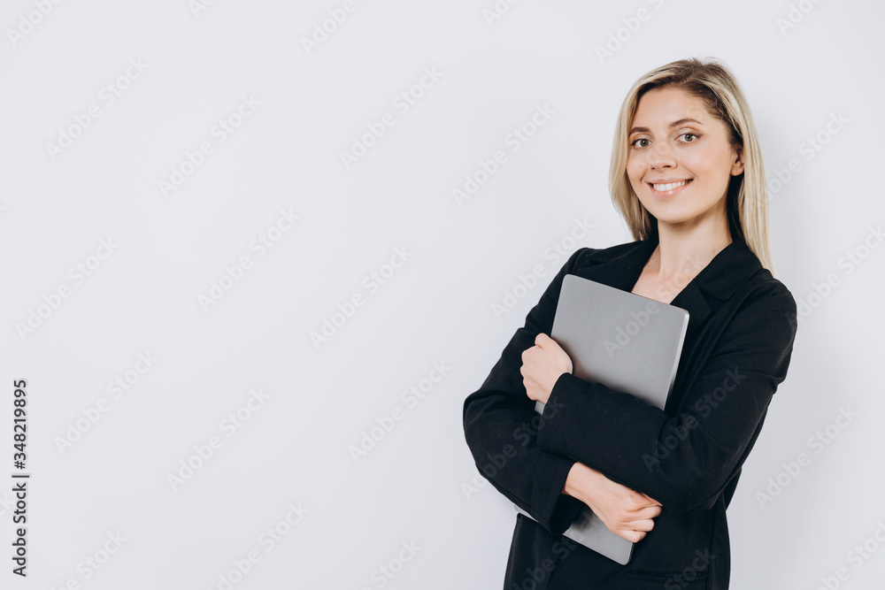 A pretty young smiling woman is standing with a notebook pc under her arm against a light background A pretty young smiling woman is standing with a notebook pc under her arm against a light background