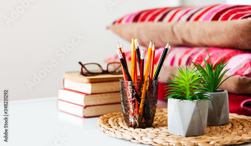 Cozy home interior decor: pencils in holder, stack of books, plants in pots on a wicker stand, pillows and plaid on a white table. Distance home education. Quarantine concept of stay home.