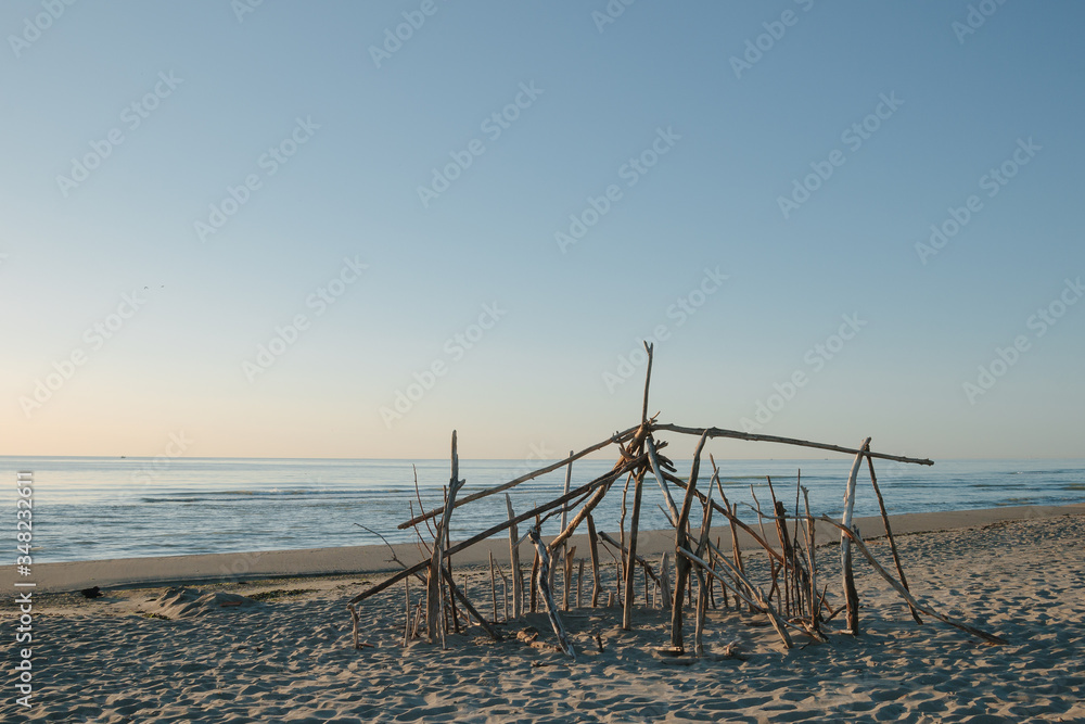 Seaside landscape in evening. Hut of branches on a beach. Relax in evening on the beach. Sunset at sea. Calm and sort seascape. Shelter of branches on the sand on the sea coast. Dry tree Seaside landscape in evening. Hut of branches on a beach. Relax in evening on the beach. Sunset at sea. Calm and sort seascape. Shelter of branches on the sand on the sea coast. Dry tree