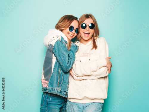 Two young beautiful brunette smiling hipster girls in trendy summer hoodie and jeans jacket clothes. Sexy carefree women posing near blue wall. Trendy and positive models having fun in sunglasses © halayalex