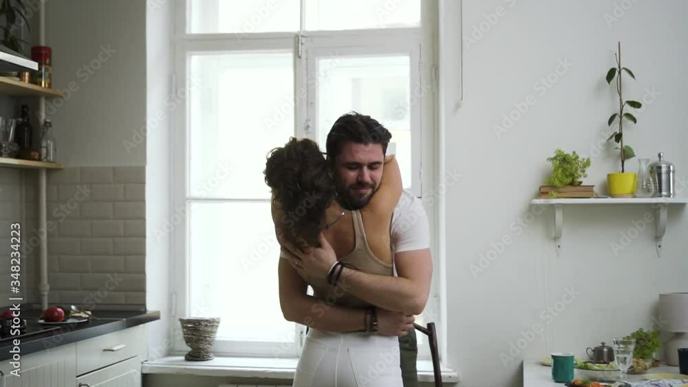 Portrait of happy american couple is dancing and cuddling in modern apartment spbd. Beautiful curly woman and bearded man move with smiles and hug tenderly, having romantic time in home kitchen. Two Portrait of happy american couple is dancing and cuddling in modern apartment spbd. Beautiful curly woman and bearded man move with smiles and hug tenderly, having romantic time in home kitchen. Two