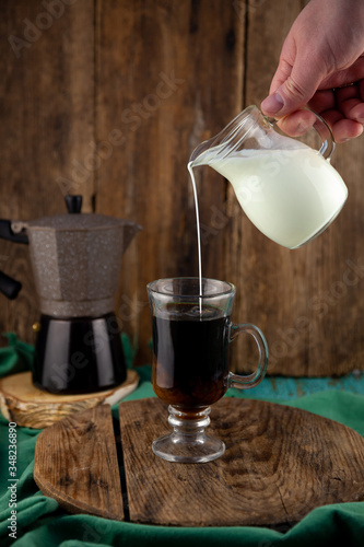 Pour milk into coffee. Make coffee with cream. Glass head in hand. A glass of cappuccino. Geyser coffee maker on a wooden table. Cook breakfast at home. Kitchen background. Carafe creamer.