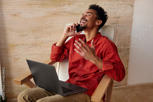 Overjoyed young pretty short haired bearded dark skinned man thowing back his head while laughing and raising emotionally hand, making phone call while sitting in chair photo