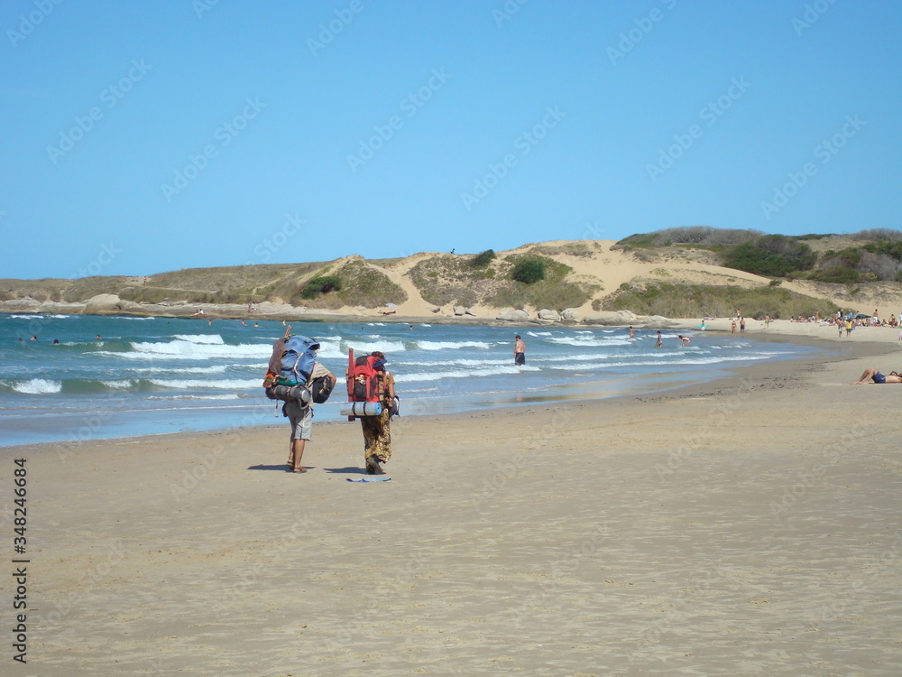 A couple of backpackers tourists on vacation goes camping and walks along the shoreline sands in Punta del diablo, Rocha, Uruguay. Atlantic ocean coast of South America. A couple of backpackers tourists on vacation goes camping and walks along the shoreline sands in Punta del diablo, Rocha, Uruguay. Atlantic ocean coast of South America.