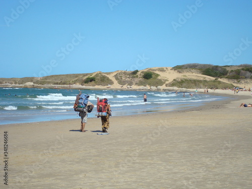 A couple of backpackers tourists on vacation goes camping and walks along the shoreline sands in Punta del diablo, Rocha, Uruguay. Atlantic ocean coast of South America.