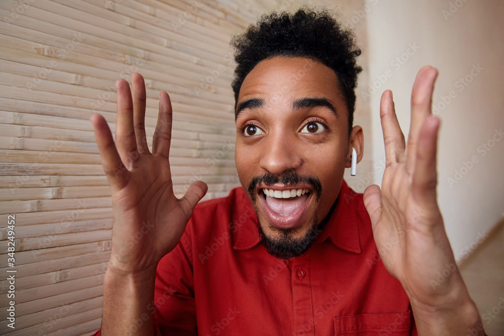 Portrait of young brown-eyed short haired bearded guy with dark skin raising emotionally hands while looking amazedly ahead with wide mouth opened, posing over home interior