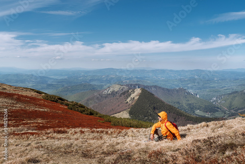 Dressed bright orange jacket backpacker with trekking poles sitting on the hill enjoying green valley at Mala Fatra mountain range,Slovakia. Active people and European hiking tourism concept image.