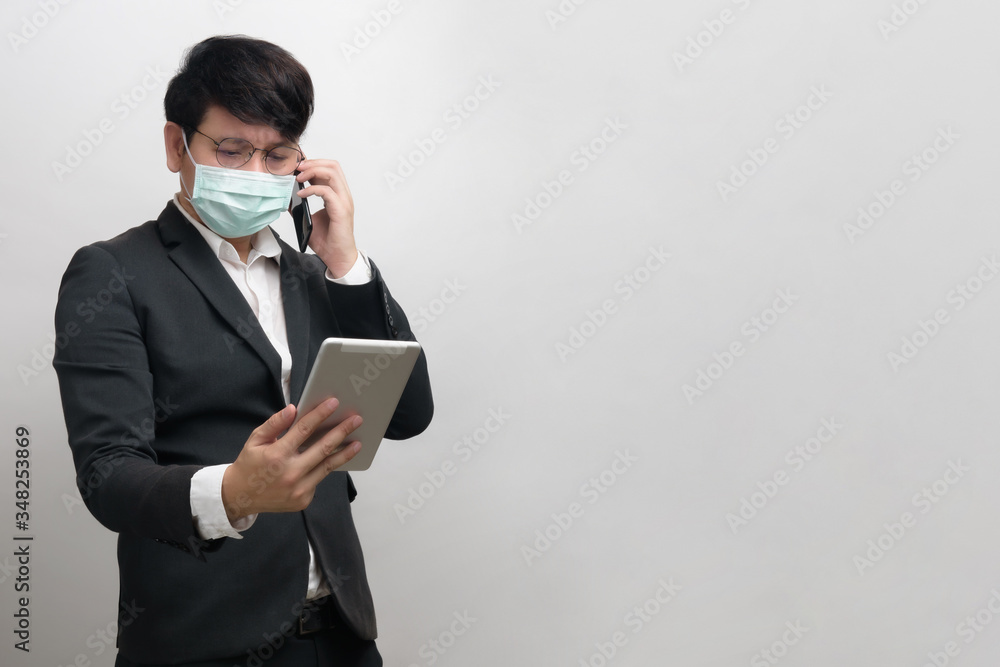 Asian businessman wearing surgical face mask in formal black suit jacket, using smart phone to talking by right hand and look at the tablet, studio lighting isolate , concept for coronavirus, covid19 Asian businessman wearing surgical face mask in formal black suit jacket, using smart phone to talking by right hand and look at the tablet, studio lighting isolate , concept for coronavirus, covid19