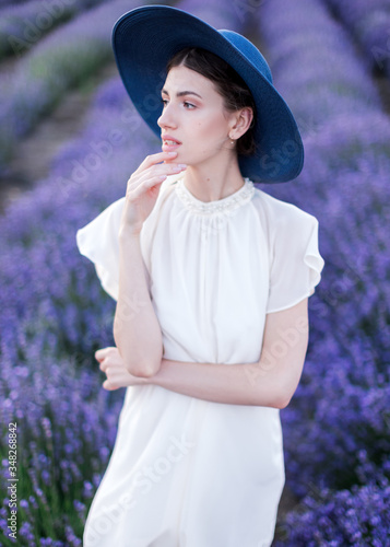 A belted portrait of a beautiful girl in a white dress and a blue hat standing in the middle of blossoming lavender fields and looking thoughtfully aside