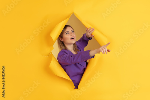 Pointing, showing. Cheerful caucasian young woman poses in torn yellow paper background, emotional and expressive. Breaking on, breakthrought. Concept of human emotions, facial expression, sales, ad. © master1305