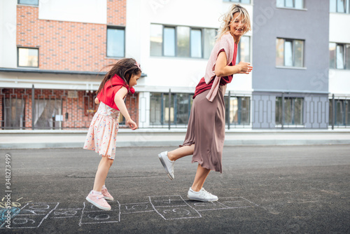 Outdoor image of happy little girl playing hopscotch with her mother on playground outdoors. Child plays with her mom oustside. Kid plays hopscotch drawn on pavement. Activities and games for children photo