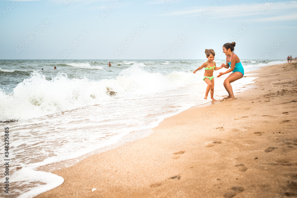 Two charming little girls in swimsuits are dancing on a sandy beach near the sea against the blue sky on a warm summer day. Concept of vacation with children