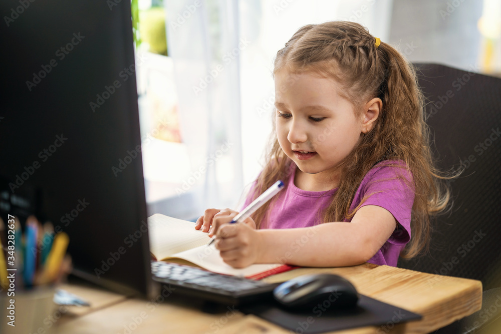 A little cute girl using computer for remote e-learning, , writes in a notebook A little cute girl using computer for remote e-learning, , writes in a notebook