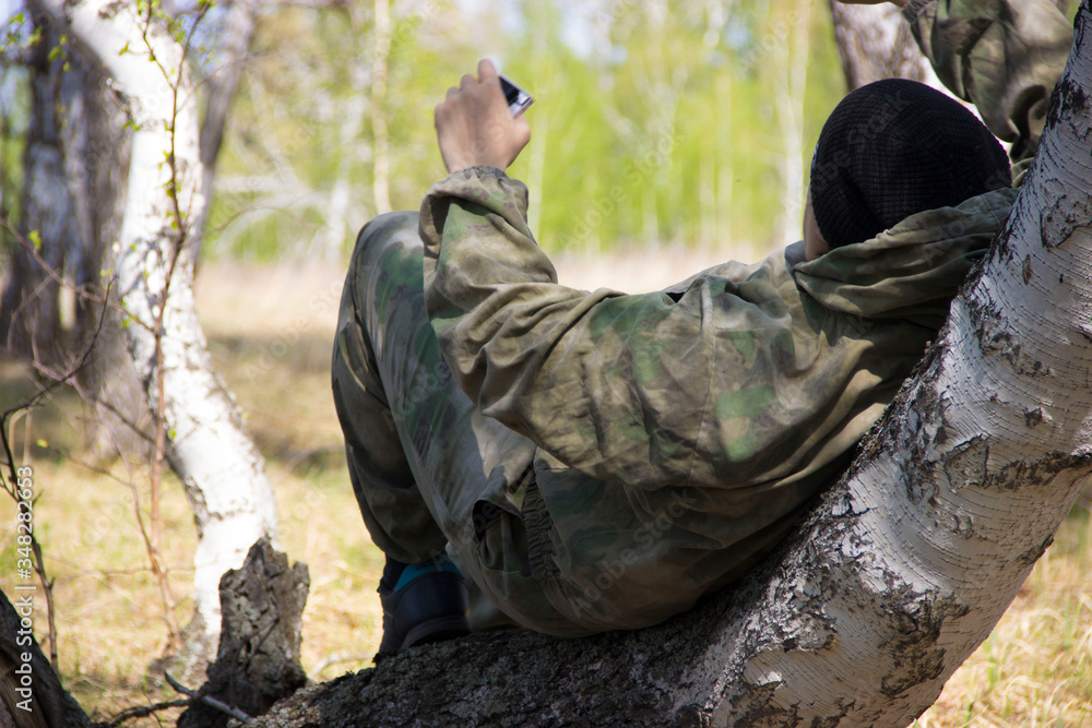 A man in camouflage clothing is resting on a curved birch.
A man in camouflage clothing is resting on a curved birch.