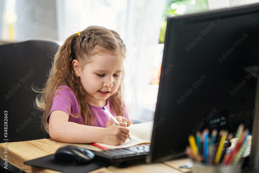 A little cute girl using computer for remote e-learning, , writes in a notebook A little cute girl using computer for remote e-learning, , writes in a notebook