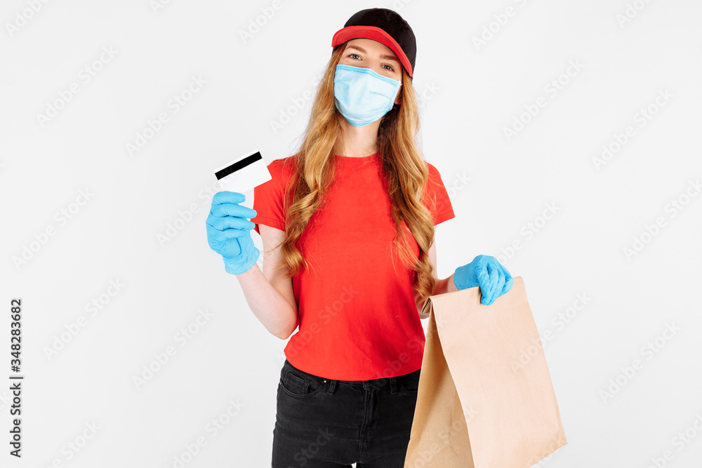 Obraz premium A courier, wearing medical rubber gloves and a medical mask, standing with a paper bag and a credit card on a white background. Online stores, payment by Bank transfer, quarantine, coronavirus