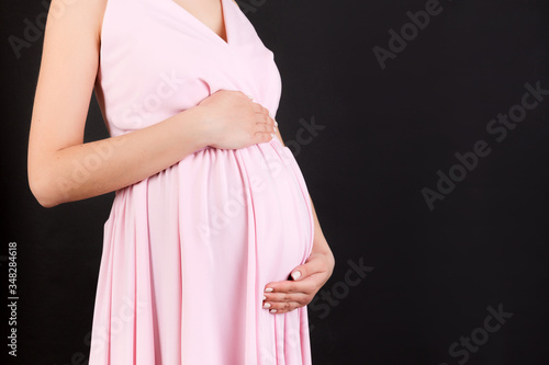 Cropped image of pregnant woman wearing pink dress. Young mother is hugging her belly expecting a baby at black background. Copy space