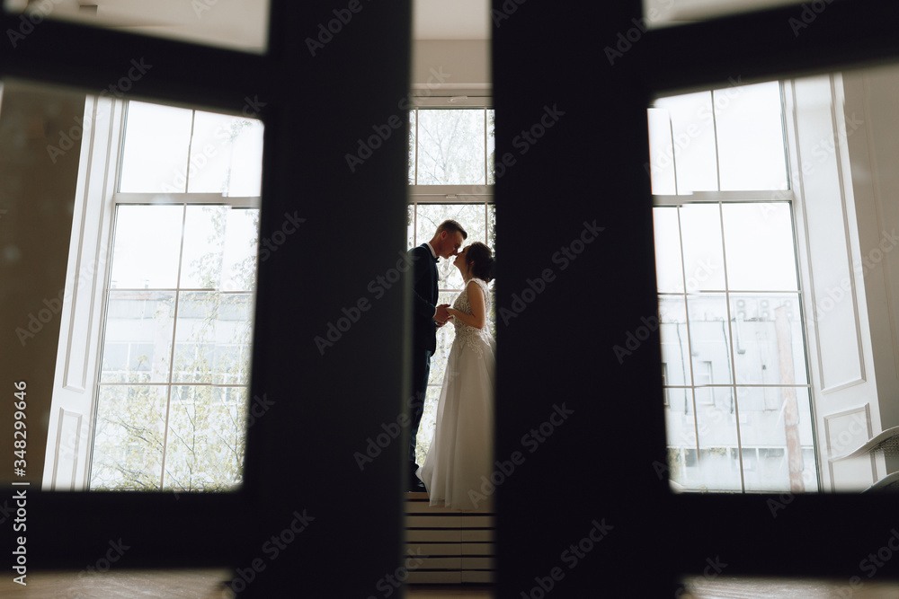 Fototapeta premium The bride and groom, European appearance. Standing at the window holding hands, looking at each other. Full height silhouette. Wedding couple.