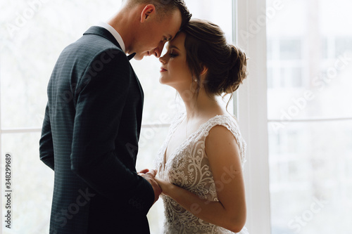 The bride and groom, European appearance. Standing at the window holding hands, looking at each other. Full height silhouette. Wedding couple.