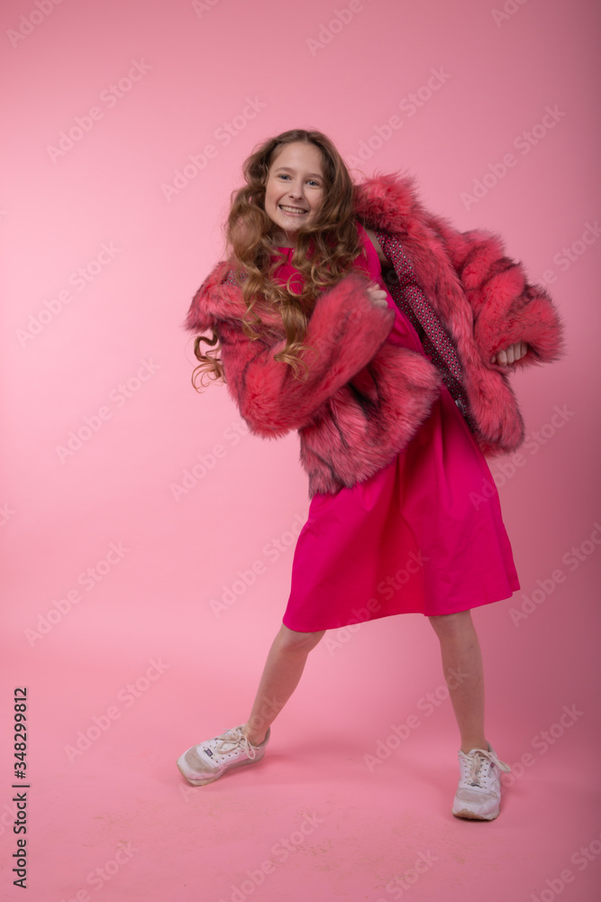 A young girl with long blond hair with curls in a fashionable fur coat with pink fur, a pink dress and white gym shoes is dancing. A young girl with long blond hair with curls in a fashionable fur coat with pink fur, a pink dress and white gym shoes is dancing.