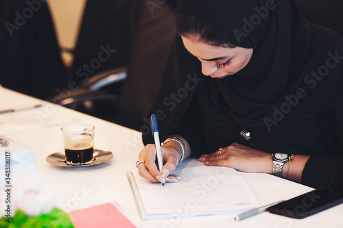 Pensive Arab woman planning working schedule writing in notebook while sitting at working place, Arabic female administrative manager making notes of information browsed on netbook.