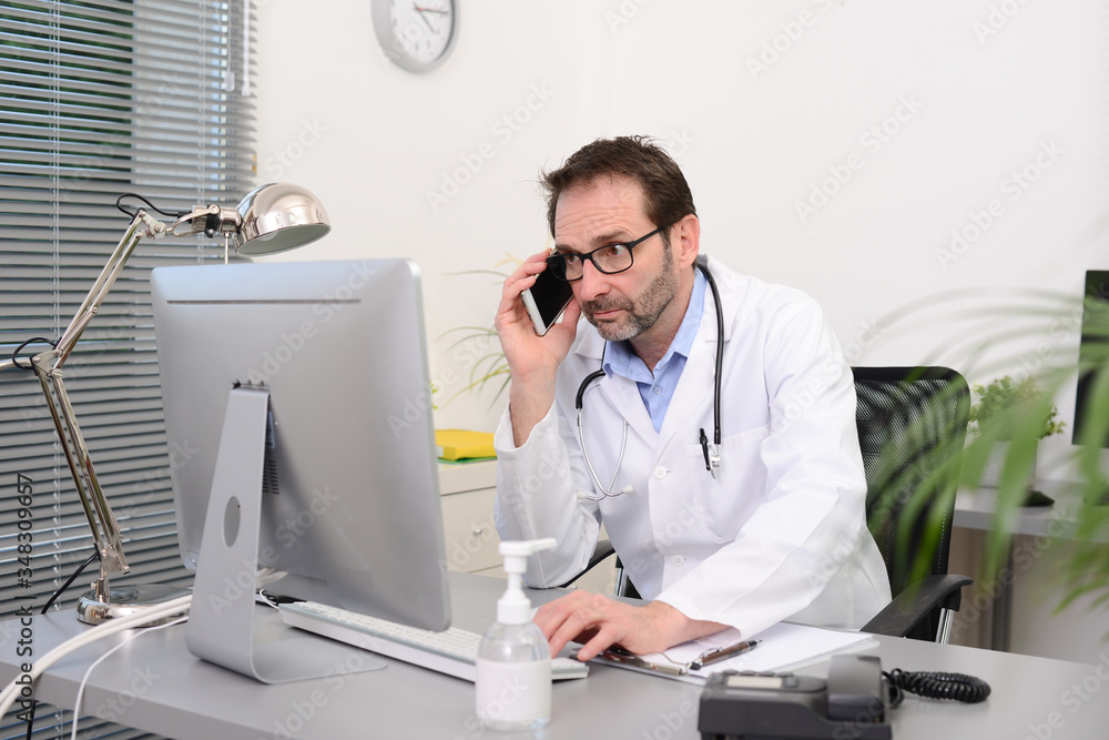 mature male doctor in medical office at desk on computer with stethoscope struggling against covid19 and coronavirus