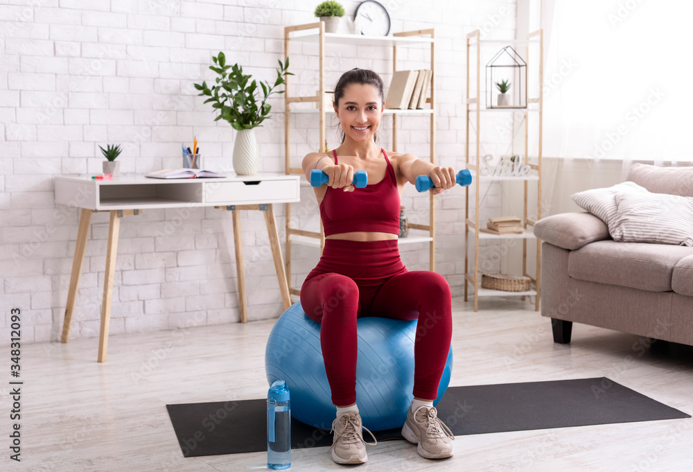 Strength training at home. Millennial girl exercising with dumbbells and fitness ball in living room