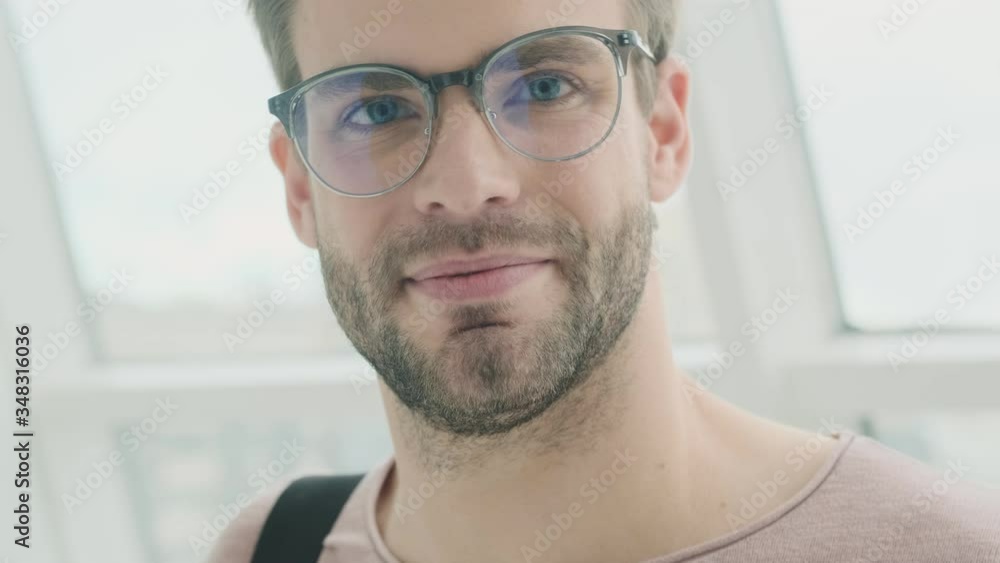 An attractive young bearded man is looking to the camera while put on his glasses while standing near the window inside An attractive young bearded man is looking to the camera while put on his glasses while standing near the window inside