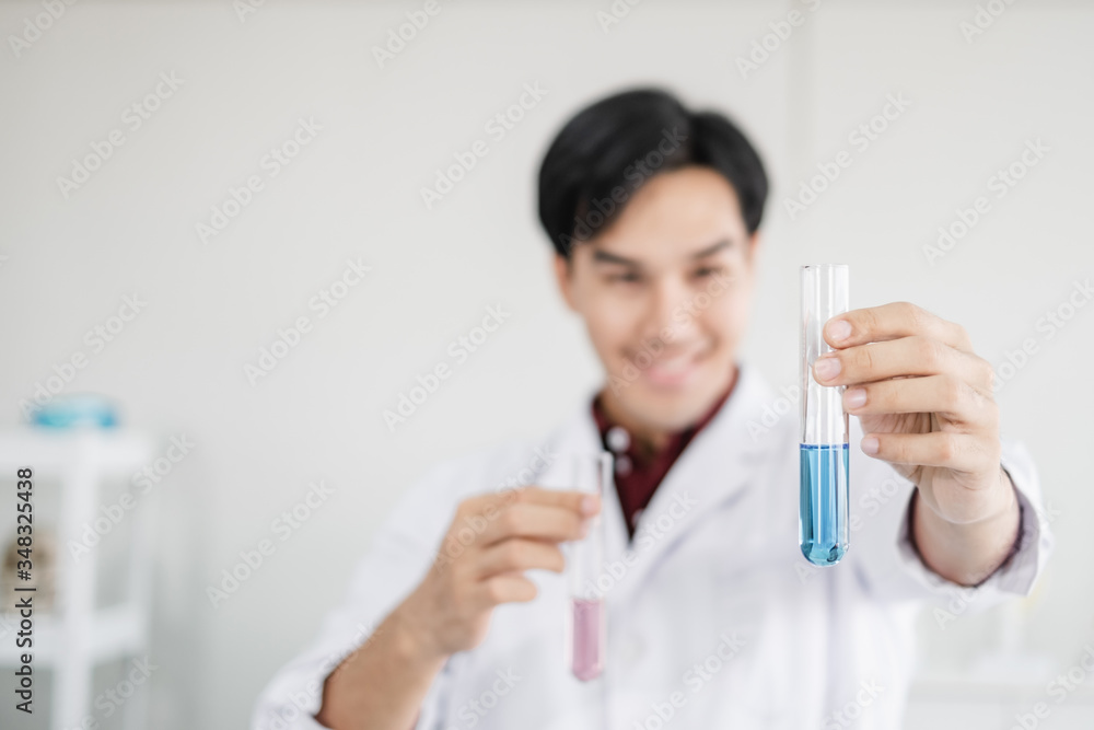 A young male scientist with black hair smiling wearing a white lab coat smiling and holding up test tubes with blue solution in a laboratory setting. Person blur in background. A young male scientist with black hair smiling wearing a white lab coat smiling and holding up test tubes with blue solution in a laboratory setting. Person blur in background.