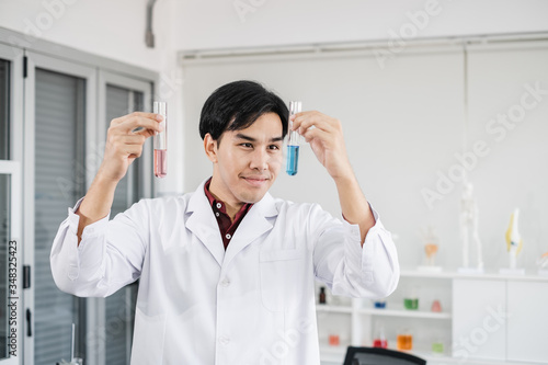 A young male scientist with black hair wearing white lab coat smiling and holding up test tubes with blue and pink solution in a laboratory setting.