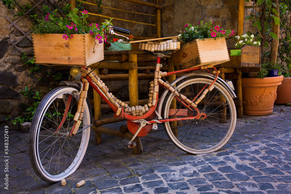 A classic red bike is decorated with wine corks and crates on a cobblestone street in Orvieto, Italy