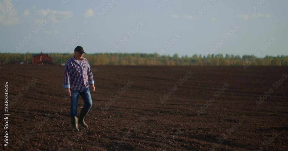 Farmer walking in idyllic,rural plowed field at sunset. Farmer wiping off sweat from forehead while walking on the field at sunset. Rubber boots for work use. A worker go with rubber boots at sunset Farmer walking in idyllic,rural plowed field at sunset. Farmer wiping off sweat from forehead while walking on the field at sunset. Rubber boots for work use. A worker go with rubber boots at sunset