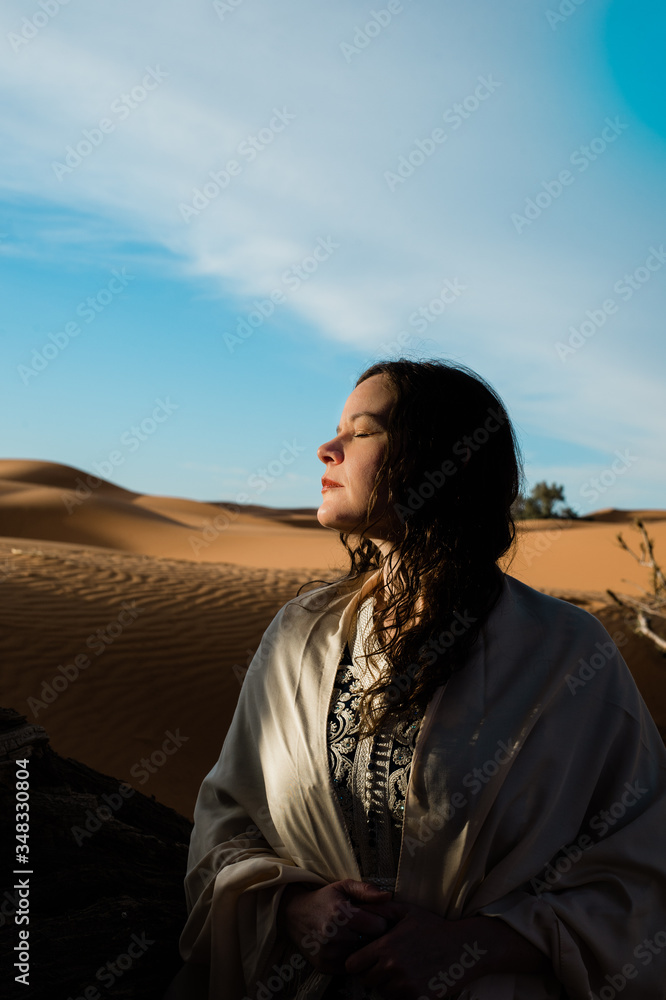 Sunlit woman in desert sand dunes with a shawl over her shoulders and a dark blue Morrocan style dress Sunlit woman in desert sand dunes with a shawl over her shoulders and a dark blue Morrocan style dress