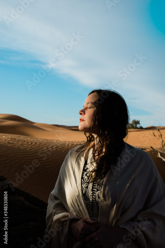 Sunlit woman in desert sand dunes with a shawl over her shoulders and a dark blue Morrocan style dress