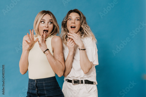 Quadro em tela Two beautiful scared blondes screaming with their hands up, dressed in casual clothes on a blue background, news about new virus