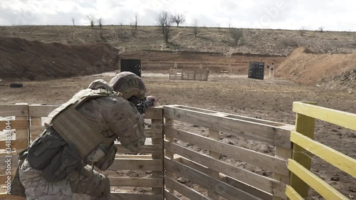 A fully equipped rookie in a camouflage shoots from an assault rifle at the training range outdoor. Bullets fly out from the gun. A unknown soldier hits the target. A shot from the back.