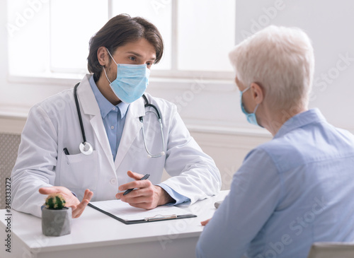 Geriatrician in medical mask talking with senior patient in office photo
