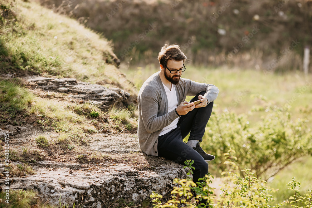 Young bearded hipster man sitting in mountains on a rock, working on the smartphone. Freelancer office in nature concept.