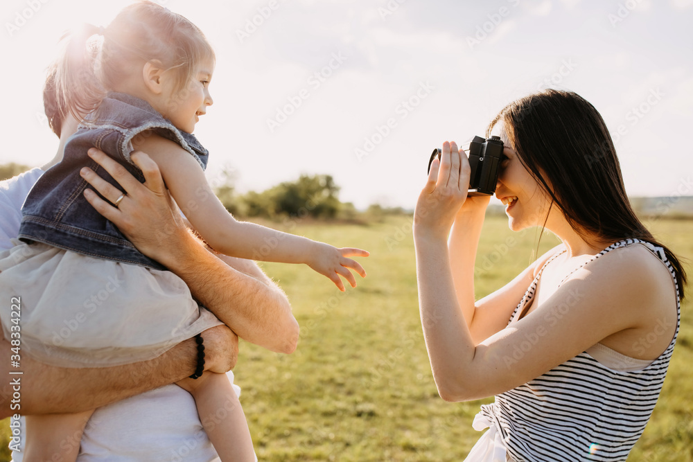 Happy family concept. Mother taking photos of father and little daughter outdoors in a field on summer day. - obrazy, fototapety, plakaty 