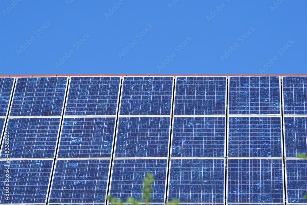 Front view of solar panels on a roof with deep blue cloudless sky in background. Photovoltaic installation of a house in daylight. Solar power system for producing electricity with sunlight. Front view of solar panels on a roof with deep blue cloudless sky in background. Photovoltaic installation of a house in daylight. Solar power system for producing electricity with sunlight.